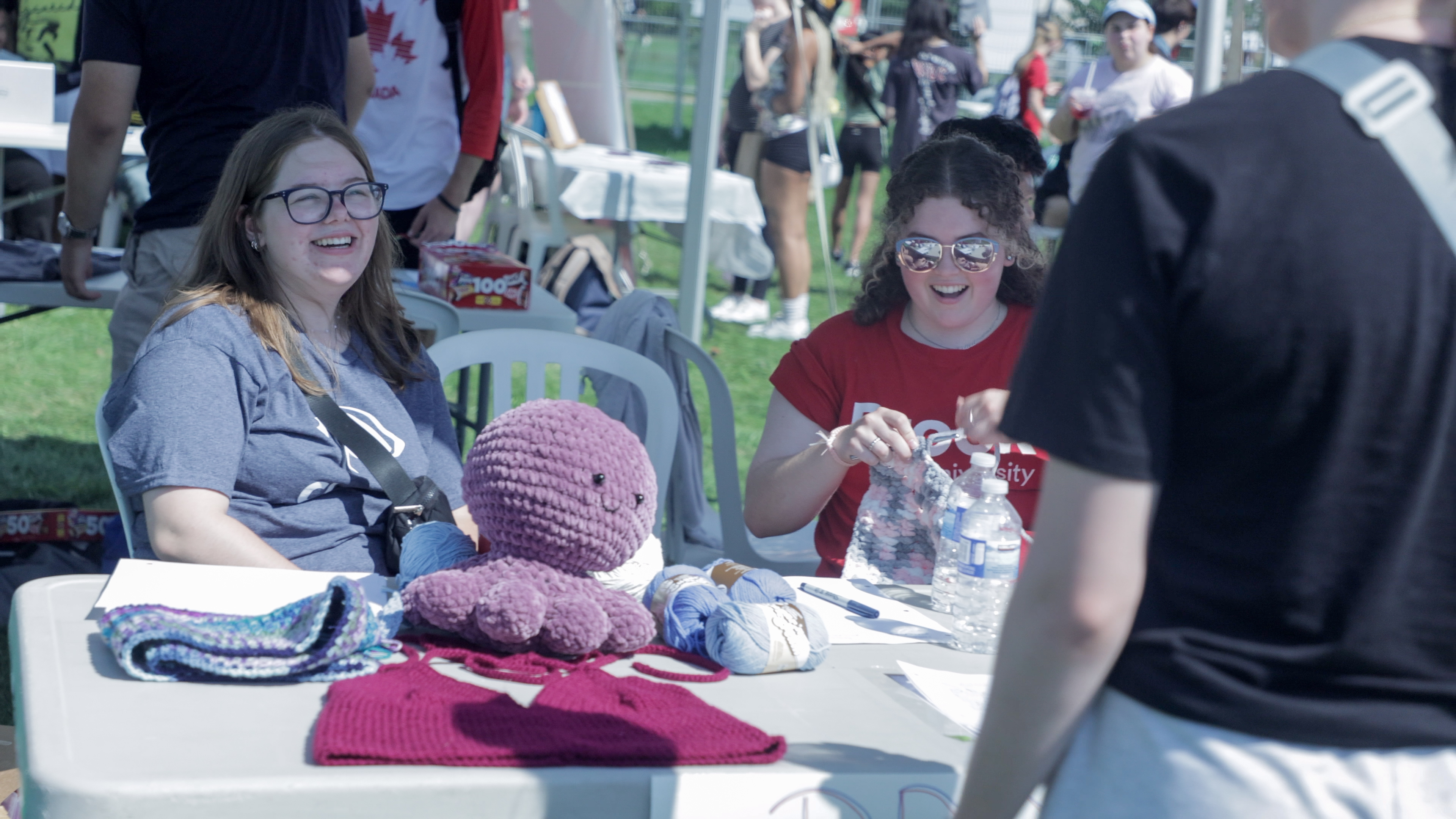 Two students during a clubs event sitting at a table, talking to another student.