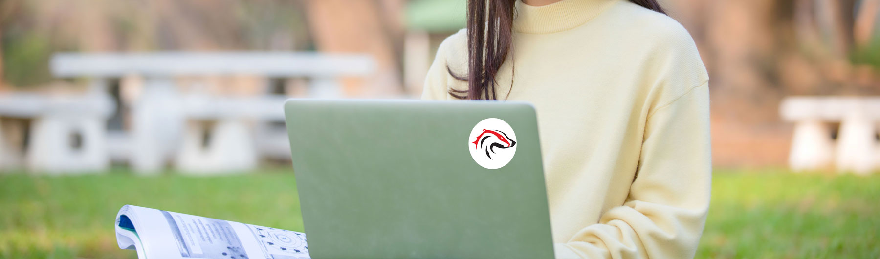 Student sitting in field smiling with laptop that has BUSU sticker
